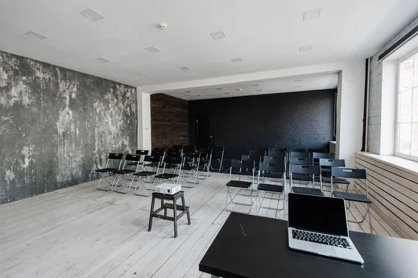 Room for lecture with a lot of dark chairs. Walls are white, loft interior. On the right there is a door. On the background there is a table with a laptop.