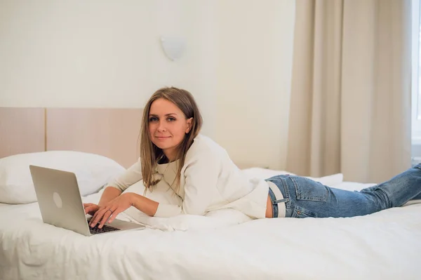 Mujer rubia atractiva pensativa usando una computadora portátil mientras se sienta en el borde de una cama en casa o en un dormitorio de hotel en vacaciones o viaje de negocios — Foto de Stock