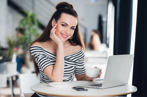 Joven hermosa mujer de negocios que trabaja en su computadora portátil en el interior, mujer adulta sonriente está utilizando un ordenador portátil en la acera cafetería, chica sonriente con computadora portátil en la cafetería . — Foto de Stock