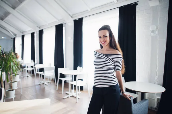 Retrato de una hermosa mujer mirando a la cámara cerca de una gran ventana. Propietario de café adulto con una sonrisa de bienvenida en el interior moderno . — Foto de Stock