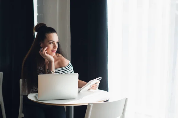 Jeune femme attrayante avec un regard réfléchi assis dans un café avec un ordinateur portable et écrire une lettre . — Photo