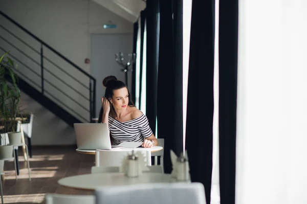 Hermosa mujer joven tomando notas en su computadora portátil y mirando a través de la ventana mientras está sentada en una mesa en un café . —  Fotos de Stock