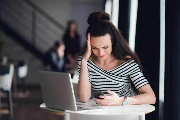 Portrait of stressed office young woman holding a cellphone in hands in a cafe, looking at the screen with cross face expression, mad at stressful texts and calls. — Stock Photo, Image