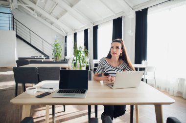 Genç kadın bir dizüstü bilgisayar kullanarak ve uzakta ile düşünceli bakışı bakarken bir cep telefonu tutan kafeterya masada oturur.