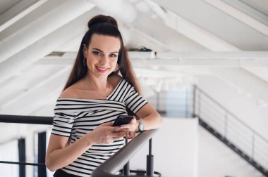 Ona telefon ile internet'te sörf işkadını gülümseyen ve kameraya bakarken gülümseyen.