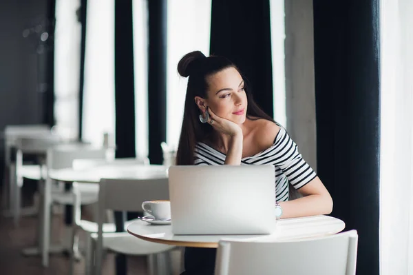 Retrato de una mujer atractiva sentada en una cafetería con su portátil y pensando en un tema mientras mira por la ventana . — Foto de Stock