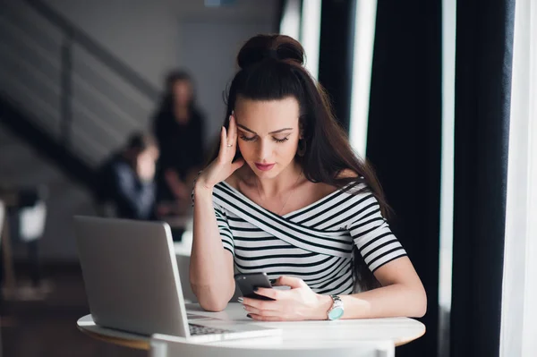 Beautiful young businesswoman using mobile phone while working with laptop in office, she is focused, trying to solve problem. Upset or disappointed expression at her face. Modern office scenery. — Stock Photo, Image