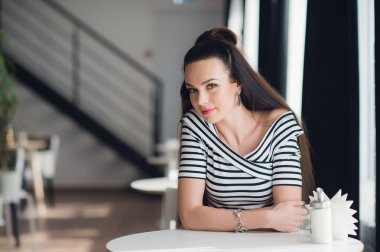 Çekici kadın esmer saçlı, seksi gülümseyen kız kapalı, bir kafede, arka plan bulanık. O güzel gülümsemesi var. Şık bir görünüm, makyaj, hafif pembe ruj, gündelik kıyafeti.