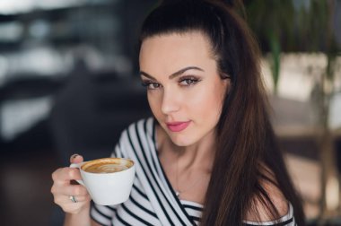 Esmer düz saç bir fincan espresso veya kapuçino ve kamera gülümseyen seyir holding ile çekici bir kadın portresi kadar yakın.