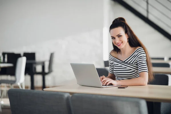 Szczęśliwy piękna freelance pracy kobieta siedząc w kawiarni z nowoczesnym wystrojem wnętrz i wpisując coś w jej laptopa. Organizowanie harmonogramu i patrząc na kamery. — Zdjęcie stockowe