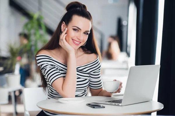 Uśmiechający się Dorosła kobieta siedząc w kawiarni z laptopa, trzymając kubek kawy i patrząc na kamery. — Zdjęcie stockowe