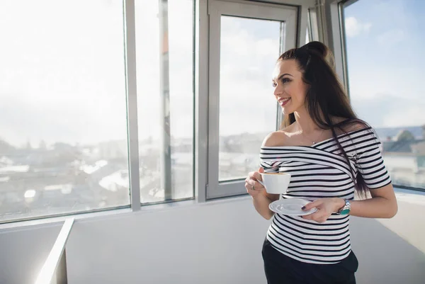 Happy woman in striped blouse smiling and standing indoors with a cup and looking away through the window.