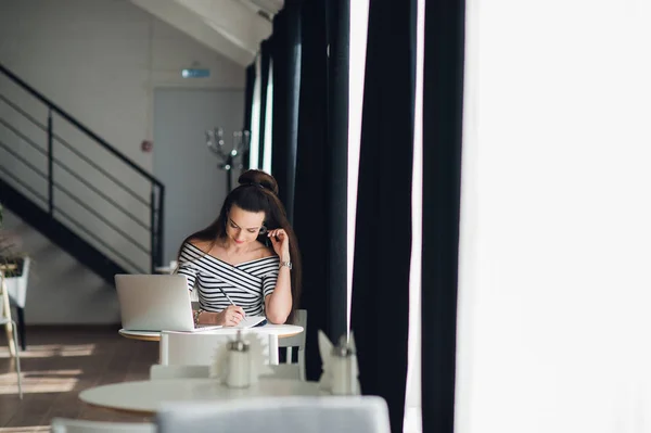 Gelukkige vrouw schrijven in een notitieblok. Volwassen vrouwtje zitten aan de tafel in de buurt van venster met een laptop. — Stockfoto