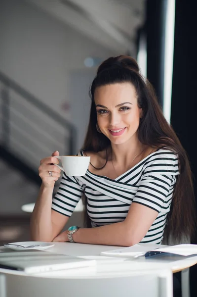 Retrato de una mujer encantadora sentada en la mesa y sosteniendo una taza de té mirando a la cámara . — Foto de Stock