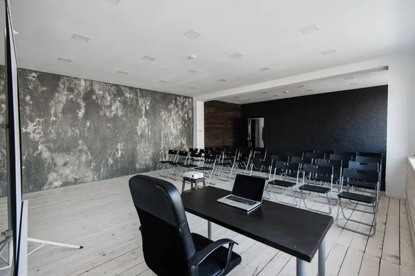 Prostor pro přednášky se spoustou tmavých židlí. Stěny jsou bílé, interiér podkroví. Na pravé straně jsou dveře. Na pozadí je tabulka s přenosným počítačem. — Stock fotografie