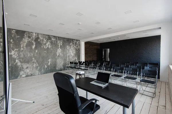 Room for lecture with a lot of dark chairs. Walls are white, loft interior. On the right there is a door. On the background there is a table with a laptop. — Stock Photo, Image