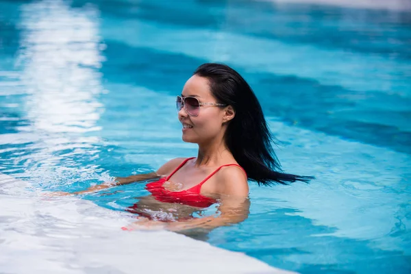 Mujer con traje de baño rojo y gafas de sol sentada en la piscina, tocando el pelo mojado — Foto de Stock
