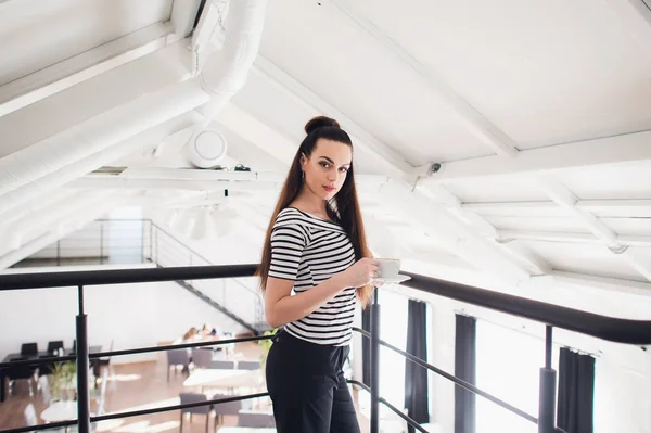 Jung und erfolgreich. attraktive junge Frau in schicker Freizeitkleidung mit Kaffeetasse und Blick in die Kamera, während sie am Fenster im Büro steht. — Stockfoto
