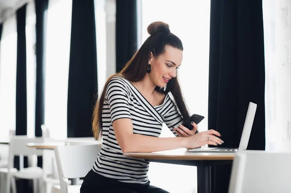 Mujer sonriente está sentada en un escritorio con una computadora portátil y mirando al sreen mientras sostiene un teléfono inteligente . —  Fotos de Stock