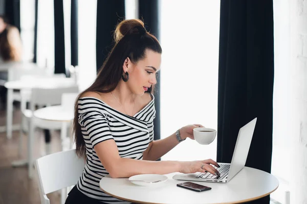 Mujer sentada cerca de la ventana en un café con un portátil. Mujer adulta sosteniendo una taza y mirando la pantalla de su computadora sorprendida . — Foto de Stock