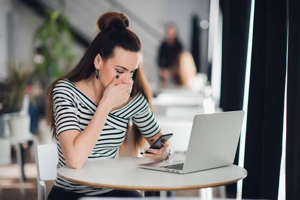 Mujer con expresión seria llena de conmoción y perplejidad está sosteniendo un teléfono inteligente mientras está sentado en un café con una computadora portátil . — Foto de Stock