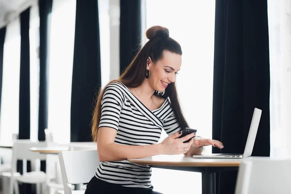 Compartir buenas noticias de negocios. Mujer joven atractiva hablando en el teléfono móvil y sonriendo mientras está sentada en su lugar de trabajo en la oficina y mirando el portátil . — Foto de Stock