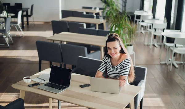 Foto de una joven hembra sonriente sentada en un café coworking mientras usa la computadora portátil chateando por teléfono. Mirando la cámara . —  Fotos de Stock