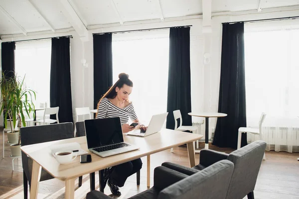 Ελκυστική γυναίκα ελεύθερος επαγγελματίας εργάζεται σε ένα ξύλινο γραφείο στις δύο laptop και smartphone ενώ περιμένουν για το co-oworker. — Φωτογραφία Αρχείου