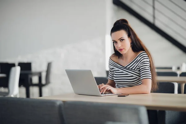 Attractive self-employed woman is working on project, using wi-fi on her laptop, sitting at wooden table with hot drink and smartphone, looking at camera with serious expression. — Stock Photo, Image