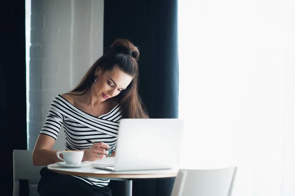 Mujer joven está sentada en el escritorio de madera cerca de la ventana y tomando notas. Adulto atractiva hembra está sentado con un ordenador portátil y una taza de café caliente, mientras que la escritura en un portátil . — Foto de Stock