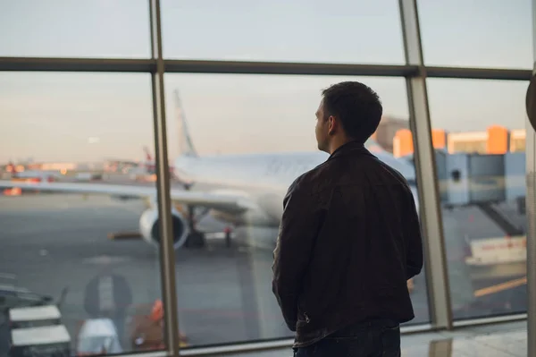 Young man is standing near window at the airport and watching plane before departure. Focus on his back — Stock Photo, Image