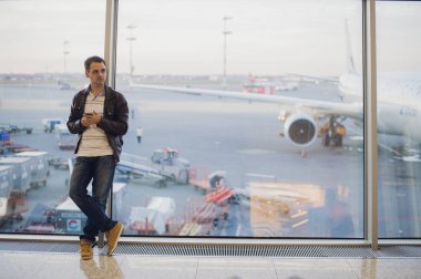 Gezgin Havaalanı terminal içinde. Cep telefonu kullanarak ve onun uçuş için bekleyen genç adam.
