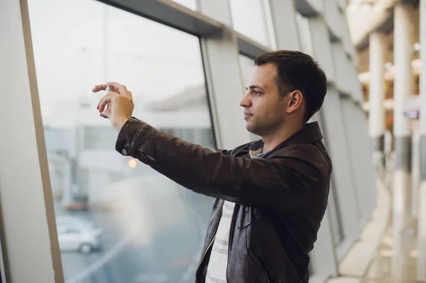 Hombre guapo de pie junto a la pared de cristal en la moderna terminal del aeropuerto, tomando foto de aviones, viajando a otros países . —  Fotos de Stock