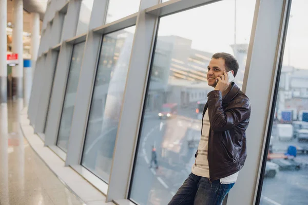 Jonge reiziger op een luchthaven, met een lederen jas praten op zijn smartphone — Stockfoto