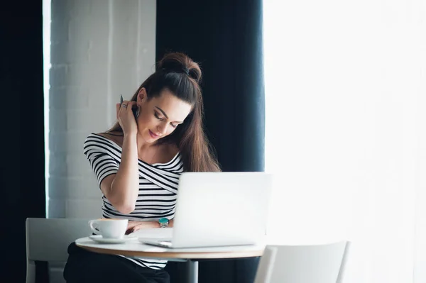 Indoor shot of charming young female with chocolate hair wearing watch on her wrist, touch face, sitting at cafe and using genic laptop computer, daylight trough window in background . — Foto de Stock