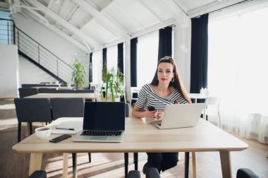 Ciddi genç iş kadını dizüstü bilgisayar ve telefon bir kafede oturan ve kameraya bakıyor.