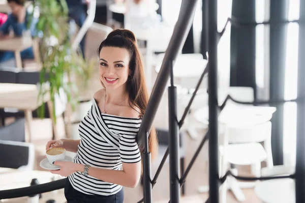 Líder de equipo seguro. Joven confiada sosteniendo una taza de café caliente en sus manos y mirando a la cámara con sonrisa mientras sus colegas trabajan en el fondo . — Foto de Stock