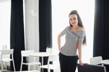 Çizgili bir gömlek veya bluz ve kot kız kameraya bakarken bir kafede duruyor. Kamera için poz ve gülümseyen sevimli seksi esmer kadın.