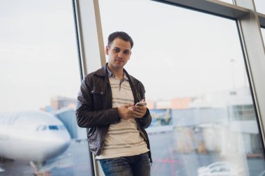 Gezgin Havaalanı terminal içinde. Cep telefonu kullanarak ve onun uçuş için bekleyen genç adam.