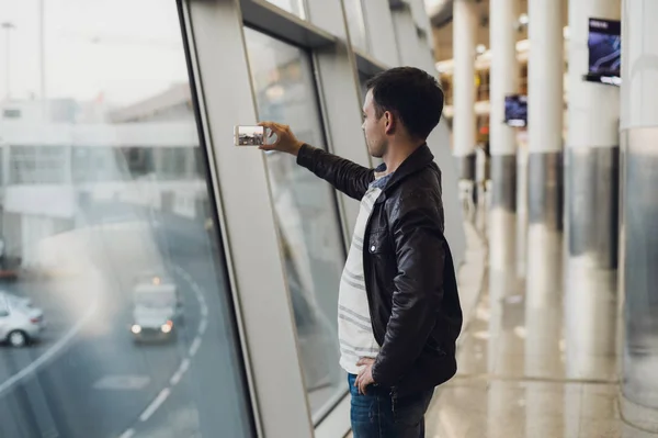 Bonito homem de pé ao lado da parede de vidro no terminal do aeroporto moderno, tirando fotos de aviões, viajando para outros países . — Fotografia de Stock