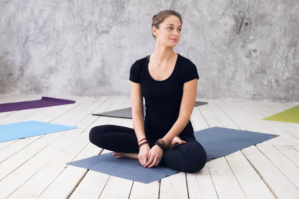 Ung attraktiv kvinna utövar yoga, sittande i Ardha Padmasana motion, halv Lotus pose, träna, klädd i svart t-shirt, byxor, meditation session på golvet vid yogaklass — Stockfoto