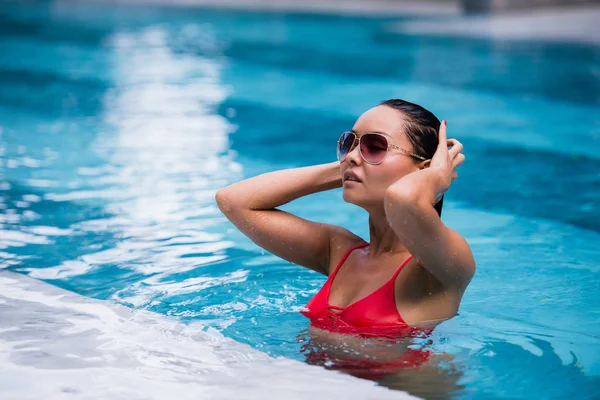 Mujer asiática en bikini naranja y lentes de sol sentados en la piscina. Retrato de moda. Mujer elegante en bikini y gafas de sol — Foto de Stock
