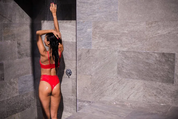 Sporty young beautiful sexy woman in a red swimsuit taking refreshing shower after swimming in the outdoor pool. Outdoor lifestyle picture on a hot sunny summer day. — Stockfoto