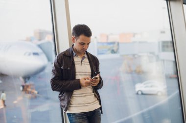 Gezgin Havaalanı terminal içinde. Cep telefonu kullanarak ve onun uçuş için bekleyen genç adam.