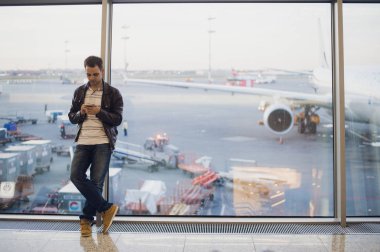 Gezgin Havaalanı terminal içinde. Cep telefonu kullanarak ve onun uçuş için bekleyen genç adam.
