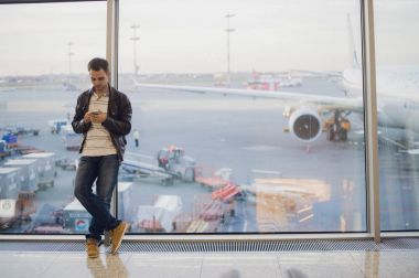 Gezgin Havaalanı terminal içinde. Cep telefonu kullanarak ve onun uçuş için bekleyen genç adam.