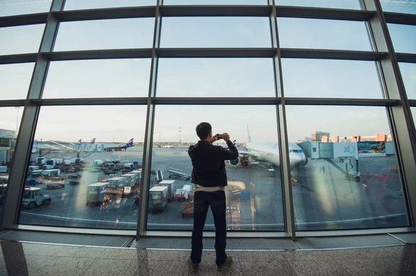 Όμορφος άνδρας στέκεται δίπλα από τον γυάλινο τοίχο στο σύγχρονο αεροδρόμιο, λήψη φωτογραφία εικόνα του αεροσκάφους αεροπλάνο, ταξιδεύουν σε άλλες countires. — Φωτογραφία Αρχείου
