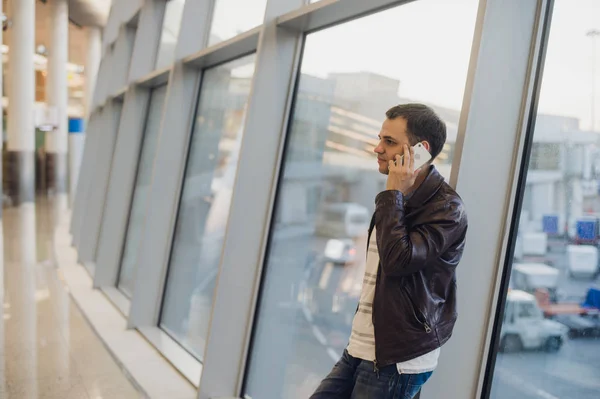 Porträtt av ung stilig person bär casual stil kläder står nära fönster i modern flygplats terminal. Resenären ringa samtal med smartphone. — Stockfoto