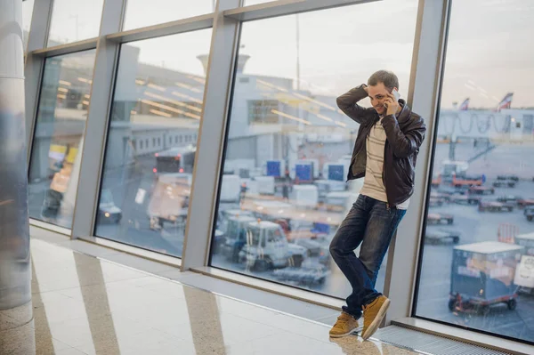Скоро буду дома. Портрет красивого молодого человека, смеющегося, разговаривающего по телефону в ожидании своего рейса в зале ожидания аэропорта — стоковое фото