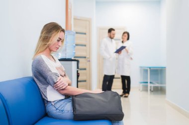 Young woman filling nervous sitting at hospital corridor waiting while two doctors talking on a backgroud clipart